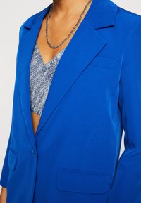 Klart blå kavaj med en strukturerad design, med en spetsig krage, två framfickor och en enkel knappstängning. Texturerat material.