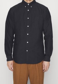 Camicia nera a maniche lunghe con colletto a punta, bottoni bianchi e taschino sul petto, abbinata a pantaloni marroni testurizzati.