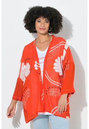 Junge Frau mit lockigem Haar, die große Creolen trägt, helle Jeans, weißes Hemd und eine lockere Kimono-Jacke mit rotem Blumenmuster.