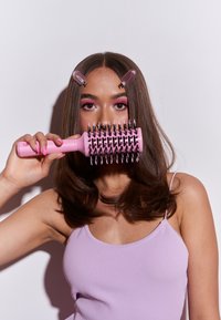 Cepillo de pelo rosa redondo con púas de metal, sostenido en una mano. La modelo tiene el cabello largo y oscuro con reflejos castaños claros y pinzas para el cabello rosas.