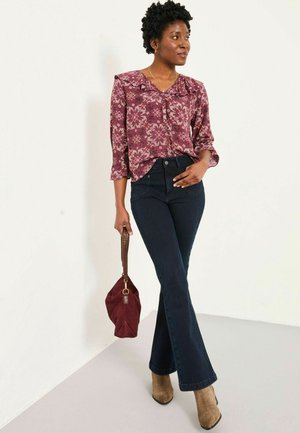Lila Bluse mit Rüschen, dunkle Schlagjeans, hellbraune High-Heel-Stiefel und eine bordeauxrote Handtasche, vor einem neutralen Hintergrund.