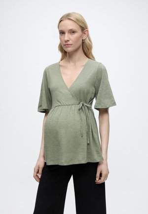 Plavolaska ženska, oblečena v svetlo zeleno nosečniško bluzo z kratkimi rokavi in črne hlače, stoji pred enostavnim belim ozadjem.