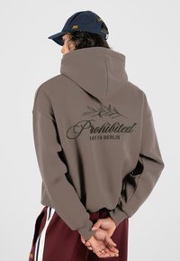 Sweat à capuche marron avec un grand texte graphique vert dans le dos, portant "Interdit 10119 BERLIN" et un motif de feuille.