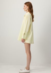 Femme aux longs cheveux roux portant une robe-chemise ample jaune pâle, des baskets blanches et des boucles d'oreilles vertes, debout de profil sur un fond uni.