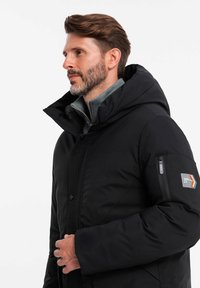 Veste doudoune noire avec capuche, dotée d'une poche zippée sur le bras gauche, de panneaux texturés et d'une finition lisse et isolante.