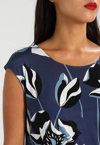 Blusa de patrón floral azul con mangas cortas tipo manga de capucha y escote ancho. El diseño incluye motivos de flores en blanco y negro sobre un fondo azul marino.