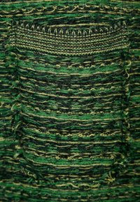 Pletená zelená látka s různými odstíny zelené a černé příze se zlatými nitěmi jako akcenty. Obsahuje texturovaný detail kapsy.