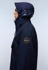 Mann in marineblauer Kapuzenjacke, der nach links blickt, mit einem Markenlogo-Patch am oberen Ärmel und sichtbaren verstellbaren Kapuzenbändern.