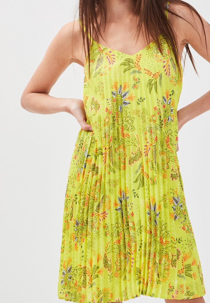 Robe florale jaune avec des bretelles spaghetti, texture plissée et décolleté en V. Présente des motifs de feuilles et de fleurs colorés sur l'ensemble.