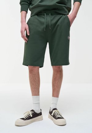 Mees, kes kannab tumerohelisi lühikesi pükse ja sobivat dressipluusi, valgeid sokke ning must-valgeid tossusid, seisab käed taskutes.