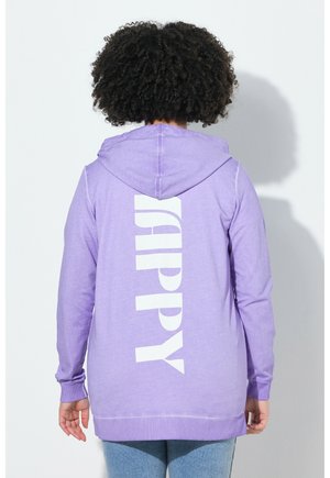 Persoon met krullend haar die een lavendelkleurige hoodie draagt met het woord "HAPPY" verticaal gedrukt op de rug, staand tegen een witte achtergrond.