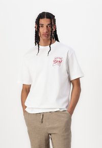White cotton t-shirt with short sleeves, featuring a red "OFF THE GRID" logo on the left chest. Worn with light brown textured pants.