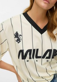Cremefarbener Jersey mit schwarzen vertikalen Streifen, versehen mit einem schwarzen Löwenemblem und dem Text "MILAN" in fetter schwarzer Schrift. Schwarzer V-Ausschnitt-Kragen.