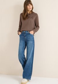 Brązowa bluza z wypukłym napisem, zestawiona z luźnymi niebieskimi dżinsami i kremowymi trampkami. Minimalistyczny design, swobodny wygląd.