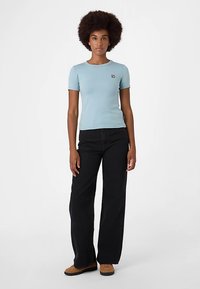 T-shirt aderente azzurro chiaro con maniche corte e un piccolo logo sul petto, abbinata a pantaloni neri a gamba larga e scarpe marroni.