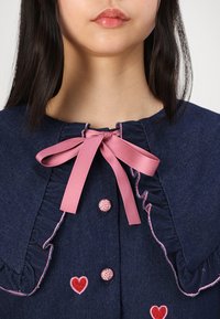 Τζιν ρούχο με ροζ ριγωτό γιακά, ταιριαστό φιόγκο και διακοσμητικά κουμπιά. Έχει κεντημένη μικρή κόκκινη καρδιά στο μπροστινό μέρος.