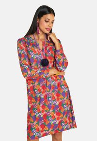 Eine Frau posiert in einem lebhaften Blumenkleid mit roten, lila und grünen Farbtönen, das durch farbenfrohen Schmuck und lange Ohrringe akzentuiert wird.