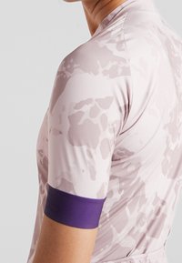 Idrottströja i ljusrosa med ett subtilt kamouflagemönster. Har en figursydd design och ett lila accentband på ärmen.