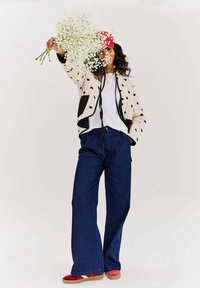 Prešívaná bunda s čiernymi srdiečkami, čierne vrecká; biela tričko; široké tmavomodré džínsy; červené tenisky; drží biele kvety.