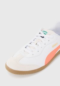 Baskets blanches avec une rayure rose-orange, orteil en daim et semelle en caoutchouc clair. Présente des lacets et un logo vert et blanc sur la languette.