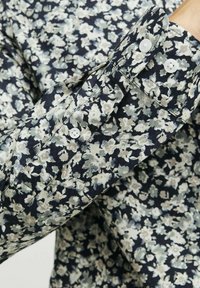 Chemise florale présentant un fond bleu marine avec des fleurs vert clair et crème, détails de poignets boutonnés et une texture de tissu lisse.