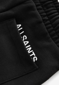 Mustast riidest lähedalt vaadatuna tasku ja valge osaliselt nähtava vertikaalselt trükitud "ALLSAINTS" tekstiga.