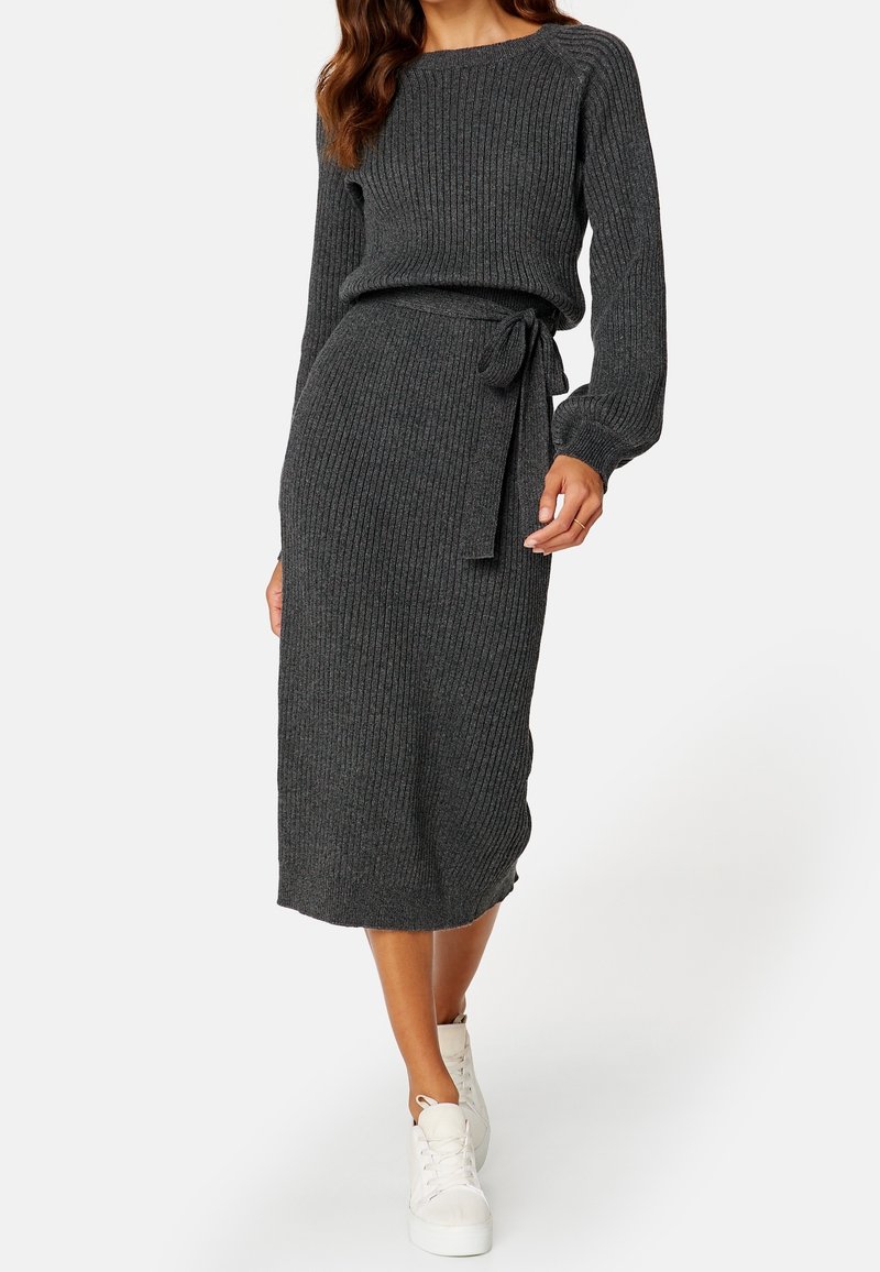 Femme portant une robe midi en maille côtelée gris foncé à manches longues, ceinture à la taille et baskets montantes blanches, debout sur un fond blanc.