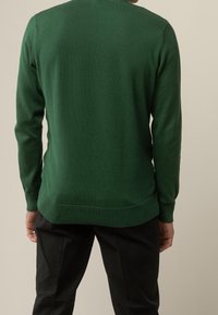 Man med mörkgrön långärmad tröja och svarta byxor, visad bakifrån mot en helt beige bakgrund.