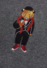 Design brodé d'un tigre en smoking avec un nœud papillon, tenant un verre, drapé dans un boa rouge, sur fond gris. Baskets rouges mises en avant.