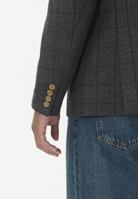 Hnědý sako s vzorem a texturovanou tkaninou, se čtyřbuttonovým manžetovým zapínáním, v kombinaci s modrými džínami. Ruka umístěná na boku.