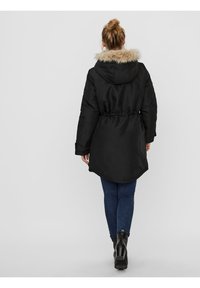Parka noire avec une capuche en fourrure, taille élastique et un dos plus long. Portée par-dessus un jean bleu et des bottines noires.