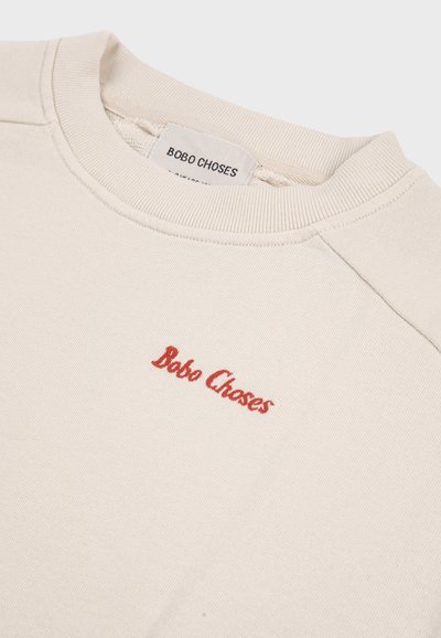 Sweat-shirt col rond couleur crème avec le texte "Bobo Choses" brodé en rouge sur le côté gauche de la poitrine et étiquette de la marque visible à l'intérieur du col.