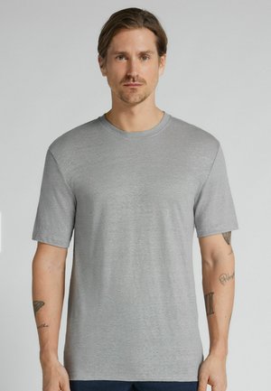 Mężczyzna z jasnobrązowymi włosami, ubrany w jednolity, szary t-shirt z krótkim rękawem, stojący na jasnoszarym tle.