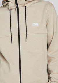 Felpa beige con zip nera e dettagli in cordino. Presenta due tasche frontali e un piccolo logo bianco nella zona sinistra del petto. Texture morbida.