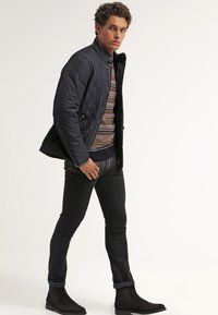 Marineblau gesteppte Jacke mit hohem Kragen, gemustertem Pullover, schwarzen Jeans und dunklen Stiefeletten. Glatte Textur und taillierte Passform.