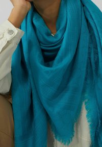 Gros plan sur une personne portant une écharpe turquoise texturée avec des bords à franges sur une veste beige clair, la main reposant près du visage.