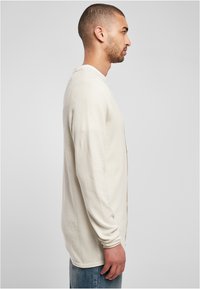 Langärmlige, hellbeige Strickjacke aus strukturiertem Stoff. Mit Knopfverschluss, runder Halslinie und kombiniert mit blauen Jeans.