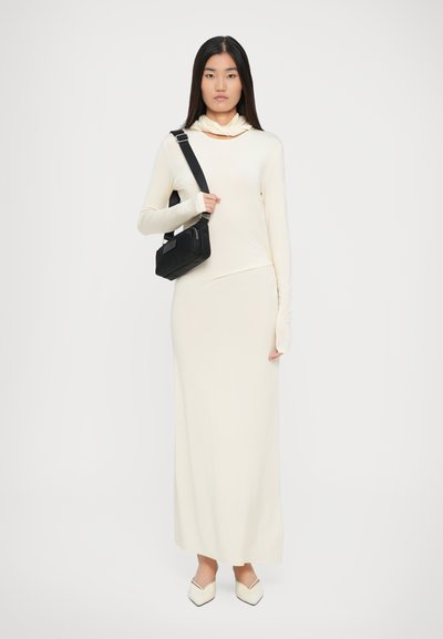 Moteris su ilgais tiesiais plaukais, vilkinti kreminės spalvos ilgomis rankovėmis suknelę ir smailiais bateliais, laiko juodą kryžminį krepšį ant balto fono.