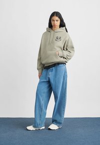 Világos zöld kapucnis pulóver elülső zsebbel és fekete logóval, laza kék farmernadrággal és fehér tornacipővel, egy kék padló háttér előtt.