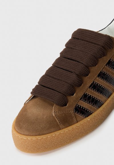Brown suede sneaker with thick brown laces, patterned black mesh sides, and a textured gum rubber sole. Curved toe and stitched accents.