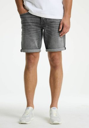 Man draagt opgerolde grijze spijkershorts, witte sneakers en een wit shirt met één hand in de zak, staand tegen een witte achtergrond.