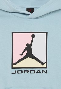 Silhueta de jogador de basquetebol a saltar com a bola dentro de um quadrado com secções rosa, branca e creme acima do texto "JORDAN" em tecido azul claro.
