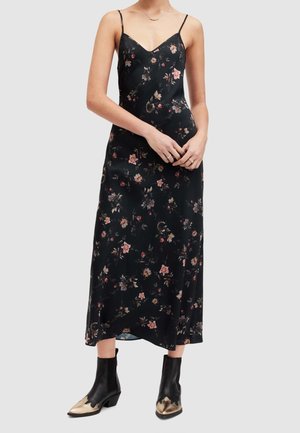 Frau trägt ein schwarzes, florales Trägerkleid mit dünnen Trägern und schwarzen Stiefeletten mit metallisch goldenen Spitzen, steht vor einem schlichten Hintergrund.
