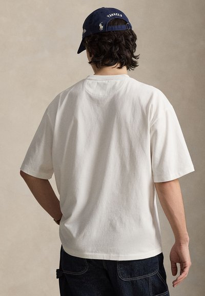 Άσπρο βαμβακερό T-shirt με κοντά μανίκια, χαλαρή εφαρμογή και στρογγυλή λαιμόκοψη. Συνδυάζεται με μπλε σκουφάκι baseball με το κείμενο και το λογότυπο "Yankees".
