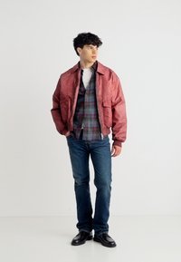 Νέος άνδρας όρθιος, φορώντας κόκκινο μπουφάν πάνω από καρό πουκάμισο, μπλε τζιν και μαύρα παπούτσια, κοιτάζοντας ελαφρώς προς το πλάι.