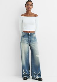 Weißes Off-Shoulder Oberteil mit langen Ärmeln, kombiniert mit hellblauen, weiten Jeans mit Spritzmustern und ausgefransten Säumen. Schwarze Schuhe.