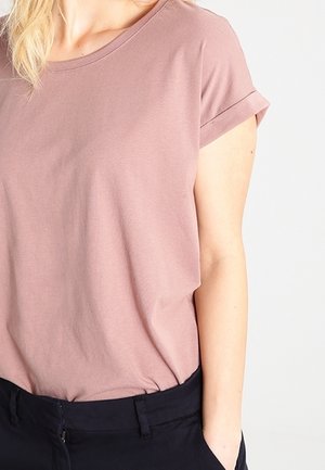 Kvinde iført en mauve kortærmet skjorte stoppet ned i sorte bukser med den ene hånd i lommen, vist fra nakke til hofter.