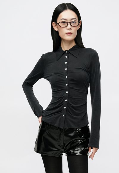 Woman with straight black hair and tortoiseshell glasses wears ruched black button-up blouse, shiny black shorts, and black tights, posing with hand on hip.
