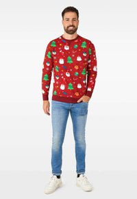 Suéter rojo con estampados festivos de muñecos de nieve, árboles y renos. Cuello y puños acanalados. Combinado con jeans de color azul claro y zapatillas blancas.