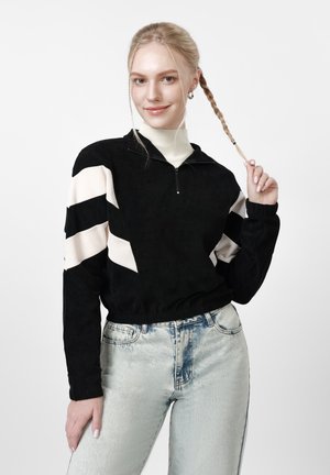 Junge Frau mit geflochtenen blonden Haaren, schwarz-weißem Langarmshirt mit Reißverschluss, hellblauen Jeans, lächelnd und hält ihren Zopf.